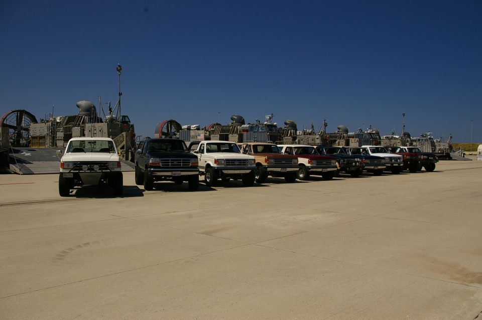 Line of Ford F-150 trucks and Broncos from the early 1990s parked on pavement with hovercraft in background