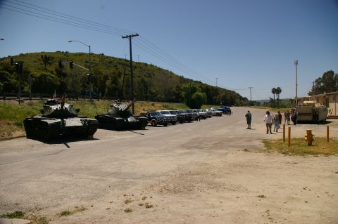 1986 Ford Bronco - A sunny day scene features two tanks parked on the side of a road, with a row of cars and a group of people nearby.