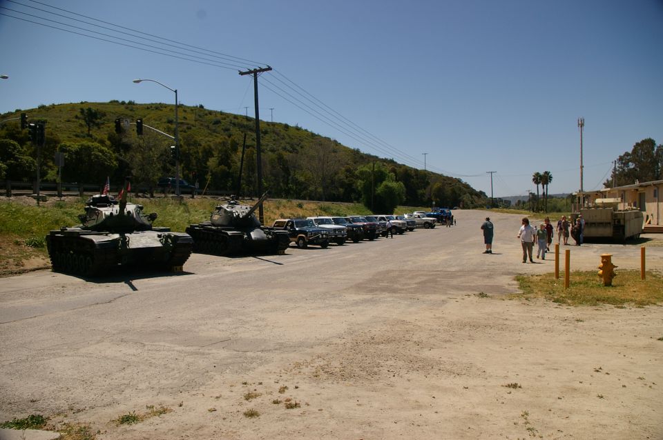 Two military tanks parked on a road next to a line of cars. People are walking near a building in the background