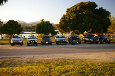 1986 Ford Bronco - A lineup of vintage SUVs and trucks, including a 1986 Ford Bronco, are parked near a tranquil lake at sunset.