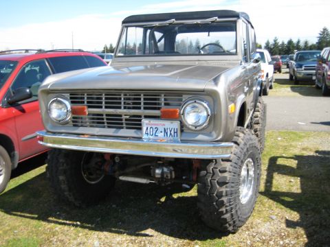 1980 Ford Bronco - This lifted 1970s Ford Bronco features a prominent grille and large off-road tires, parked next to a red SUV.