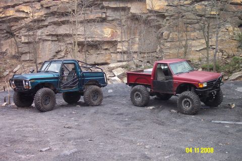 1992 Ford Bronco - Two modified trucks sit parked on a dirt lot with a rocky cliff face in the background.