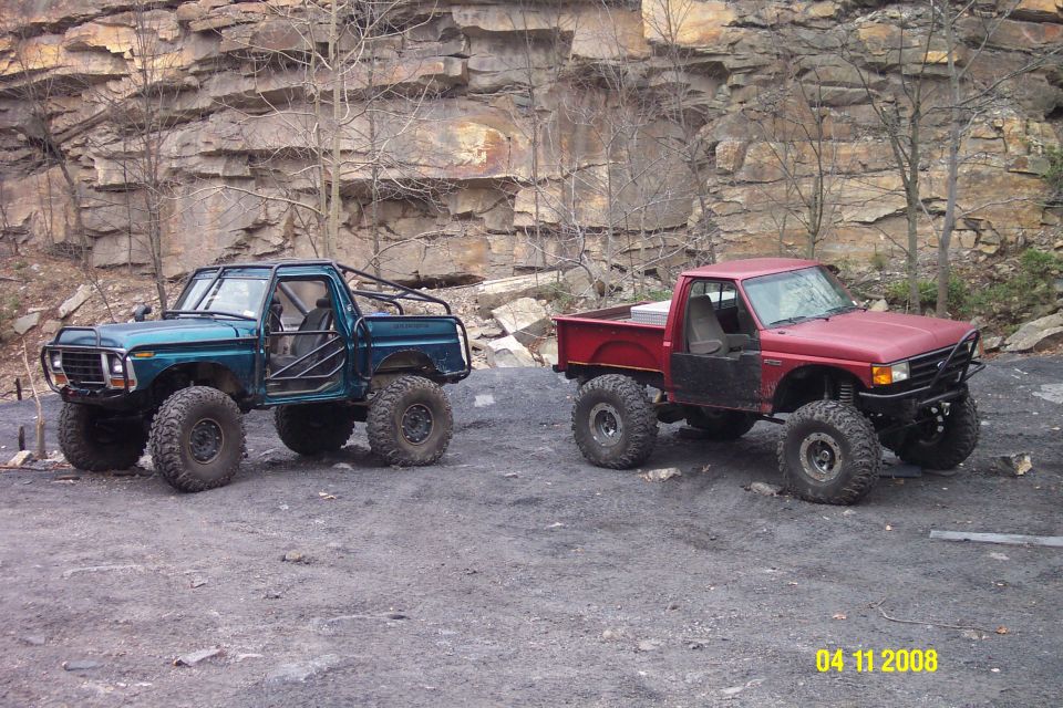 Green off-road truck with roll cage and red pickup truck with off-road tires parked on dirt near rocky cliff