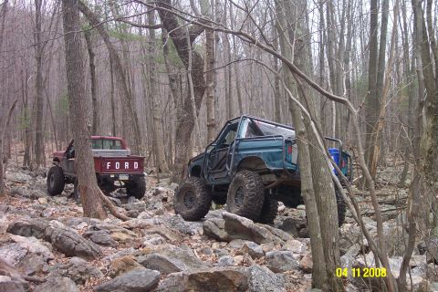 1992 Ford Bronco - Two modified off-road vehicles navigate a challenging rocky terrain in a wooded area.