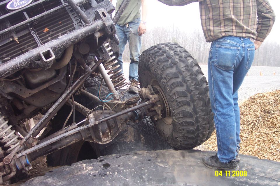 Ford Bronco front end with large off-road tires, lifted suspension, and custom roll cage. The vehicle is tilted