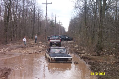 1992 Ford Bronco - Several off-road vehicles navigate a muddy trail through a wooded area with power lines overhead.