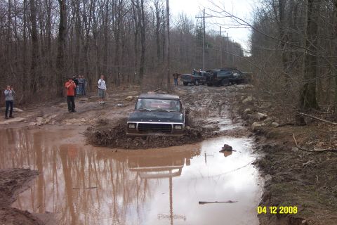1992 Ford Bronco - A black Ford F-Series truck is stuck in a muddy puddle on a wooded trail.