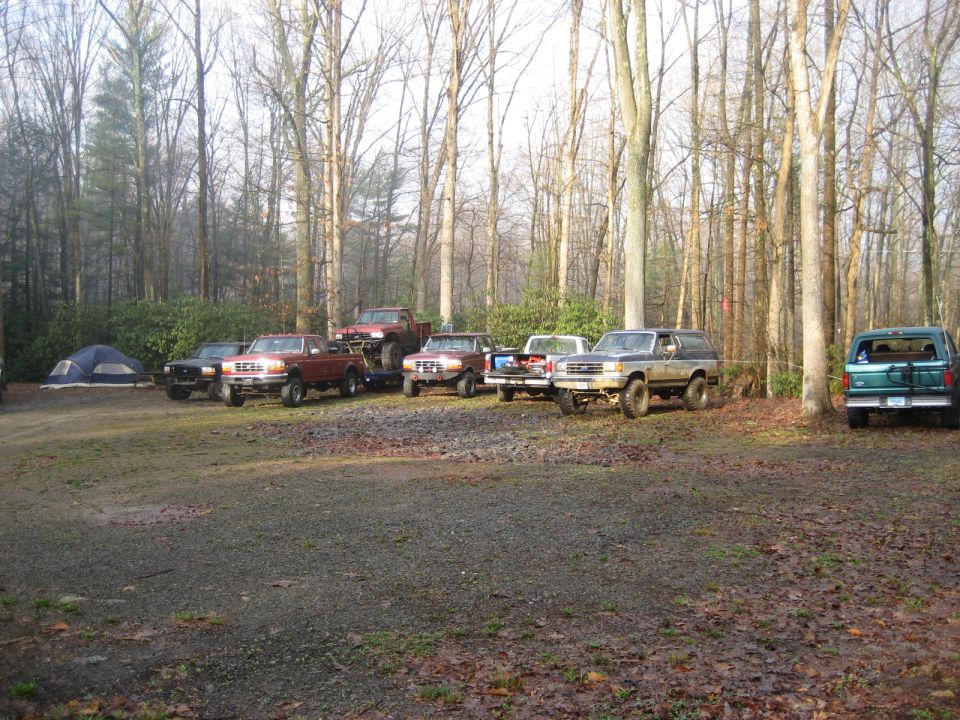 Several Ford trucks and a Bronco parked in a wooded area with tents