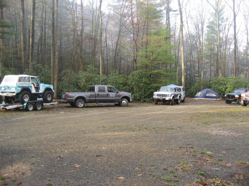 Several trucks and a trailer with a Jeep-like vehicle are parked in a wooded area with tents set up