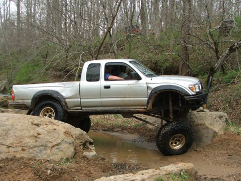 1996 Ford Bronco - A modified pickup truck drives through a muddy creek bed surrounded by trees.
