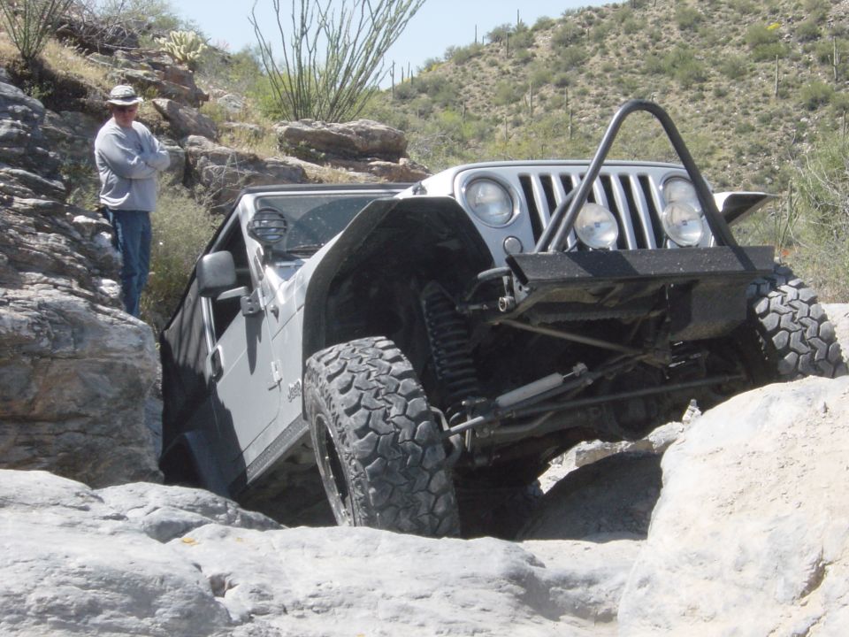 Man in hat watches a silver Jeep Wrangler navigate a rocky trail