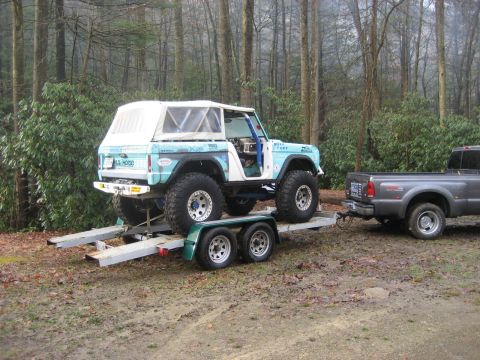 1991 Ford Bronco - A modified Ford Bronco is loaded onto a trailer, ready to be towed by a pickup truck through a wooded area.