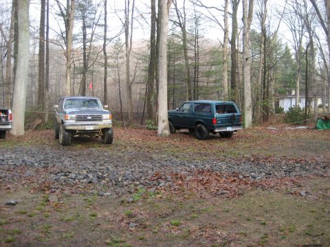 1991 Ford Bronco - Two Ford Broncos are parked in a wooded area with a house in the background.