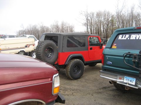 1991 Ford Bronco - A red Jeep Wrangler is parked next to a green Ford Bronco, with a trailer in the background.