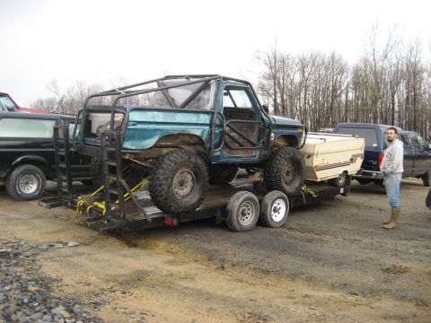 1991 Ford Bronco - Modified teal SUV with a roll cage is transported on a trailer, accompanied by other vehicles and a man.