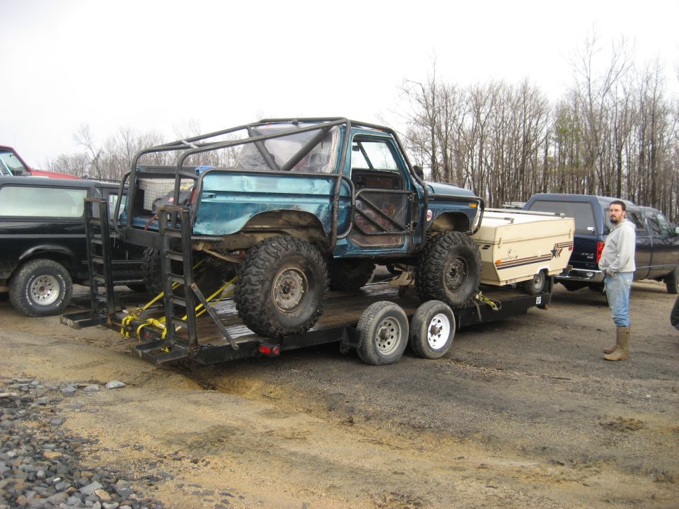 Teal off-road SUV with roll cage on a trailer, large tires, open doors, towing a small camper