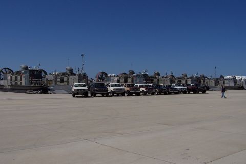 1990 Ford Bronco - A line of SUVs, including a white 1990 Ford Bronco, are parked on a tarmac with hovercraft in the background under a clear blue sky.