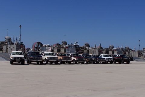 1990 Ford Bronco - A convoy of 1990 Ford Broncos and trucks are lined up on a tarmac, with hovercraft vessels visible in the distance.