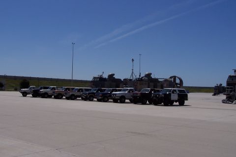 1990 Ford Bronco - A group of SUVs and trucks are parked in formation on a sunny day, with large, unusual vehicles in the distance.