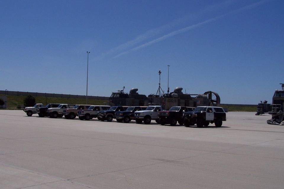 Several SUVs and trucks parked in a line on a paved lot with large, industrial vehicles in the background
