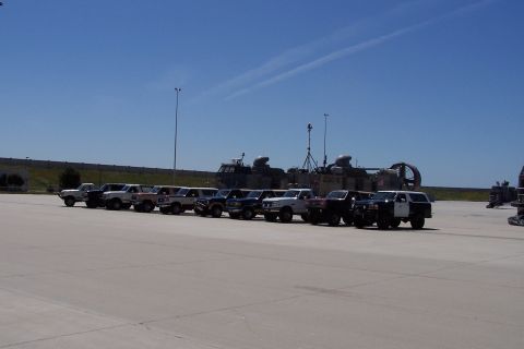 1990 Ford Bronco - Several Ford trucks and SUVs are parked in a line on a sunny day, with large military vehicles in the background.