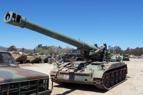 1990 Ford Bronco - A military self-propelled howitzer is parked next to a pickup truck, with other military vehicles in the background.