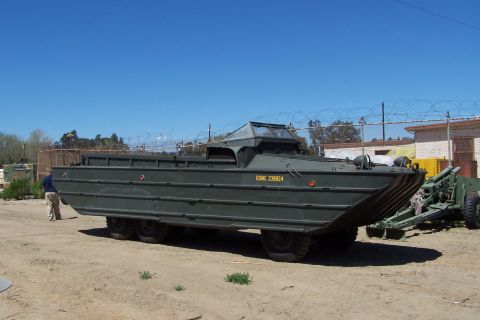 1990 Ford Bronco - A military amphibious vehicle is parked outdoors next to a cannon under a clear blue sky.