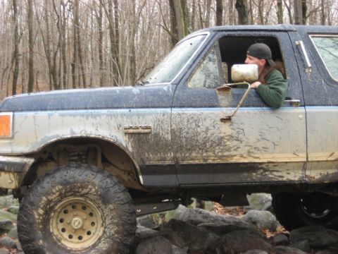 1991 Ford Bronco - A mud-splattered Ford Bronco navigates a rocky trail in a wooded area.