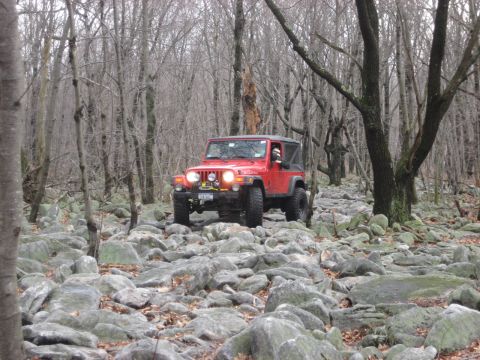 1991 Ford Bronco - A red Jeep Wrangler navigates a challenging rocky path through a bare, wooded area.
