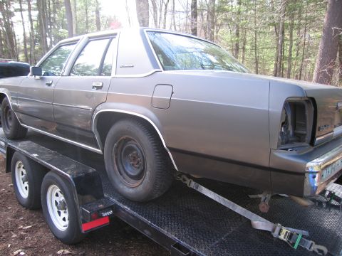 1991 Ford LTD Crown Victoria - A gray sedan is being transported on a trailer, showing its rear end with a missing taillight.