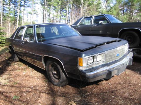 1991 Ford LTD Crown Victoria - This dark sedan, possibly a 1990s Ford Crown Victoria, is parked outdoors with its hood open and a flat tire. Another similar vehicle is vi…