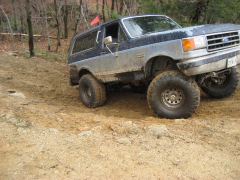 1991 Ford Bronco - This lifted Ford Bronco navigates a challenging off-road trail with muddy tires and a red flag on the roof.