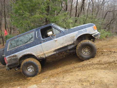 1991 Ford Bronco - A muddy Ford Bronco navigates an off-road trail with trees in the background.