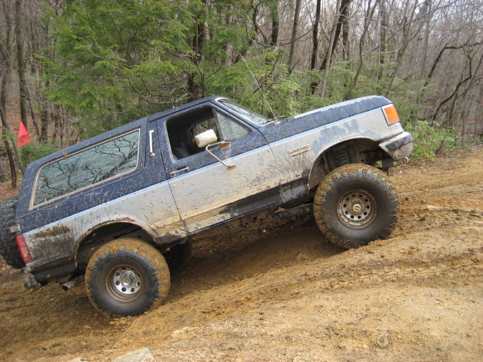 Mud-covered blue and silver Ford Bronco climbing a dirt hill, spare tire on rear door