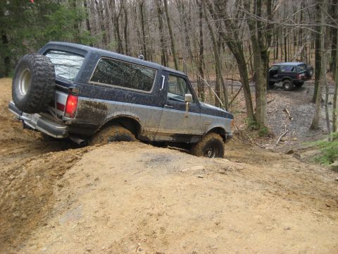 1991 Ford Bronco - Two Ford Broncos navigate a muddy off-road trail in a wooded area.