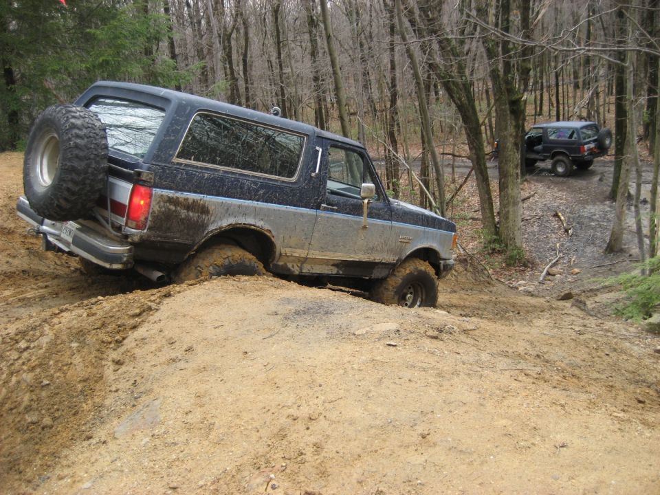1991 Ford Bronco struggling through mud on a wooded trail, another Bronco visible in the background