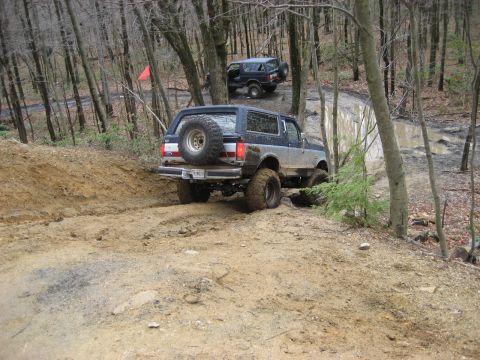1991 Ford Bronco - Two Ford Broncos navigate a challenging muddy off-road course surrounded by dense woods.