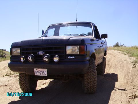 1990 Ford Bronco - This black Ford truck, seen from the front, has KC lights and a 4x4 badge, driving on a dirt path.