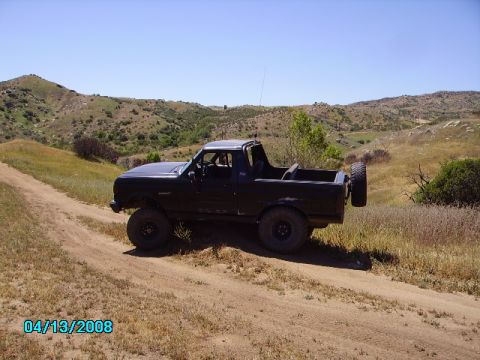 1990 Ford Bronco - A black Ford Bronco is shown on a dirt path with dry grass and hills under a clear blue sky.