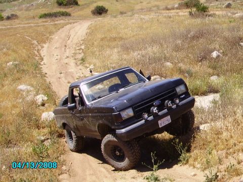 1990 Ford Bronco - A black 1990 Ford Bronco navigates a dusty off-road trail with large tires and auxiliary lights.