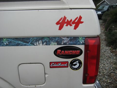 1980 Ford Bronco - Tailgate of a white pickup truck various aftermarket performance and off-road stickers.
