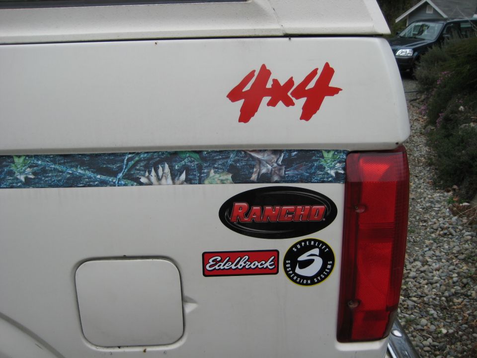 White pickup truck tailgate with 4x4, Rancho, Edelbrock, and Superlift Suspension Systems stickers. Another vehicle is visible in the background