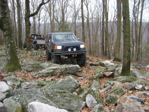 1991 Ford Bronco - This lifted Ford Bronco is tackling a challenging rocky path in a wooded area, with another vehicle following behind.