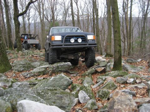 1991 Ford Bronco - A dark blue Ford Bronco navigates a challenging rocky terrain in a wooded area, with another off-road vehicle visible in the background.