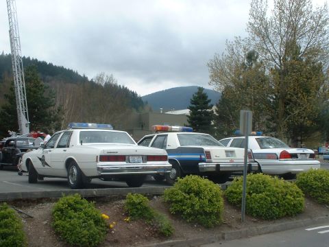 1987 Ford Crown Victoria LTD - Several police cars are parked in a lot with a fire ladder truck visible to the left.
