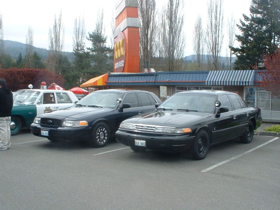 Two black police sedans parked side-by-side in a parking lot; one white and green police car is partially visible to the left