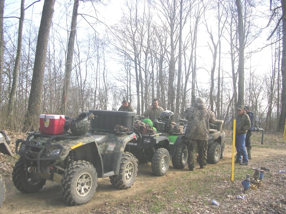 Camo-wrapped ATV with cooler and cargo box parked with other quads on a wooded dirt trail