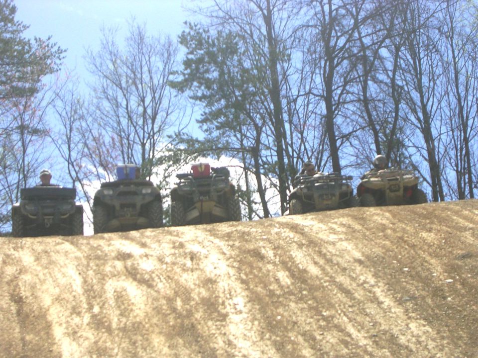 Five muddy ATVs with riders lined up on a dirt ridge against bare trees and blue sky