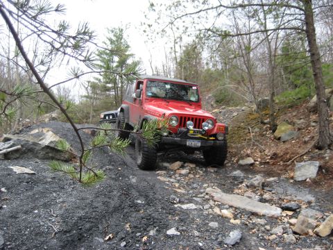 1991 Ford Bronco - Two off-road vehicles navigate a challenging, debris-strewn path through a wooded area.