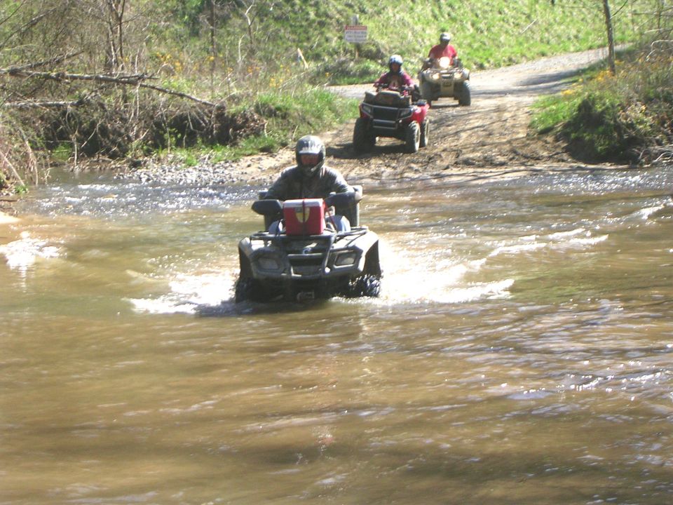 Rider on utility ATV fording a muddy creek with two quads waiting on the trail